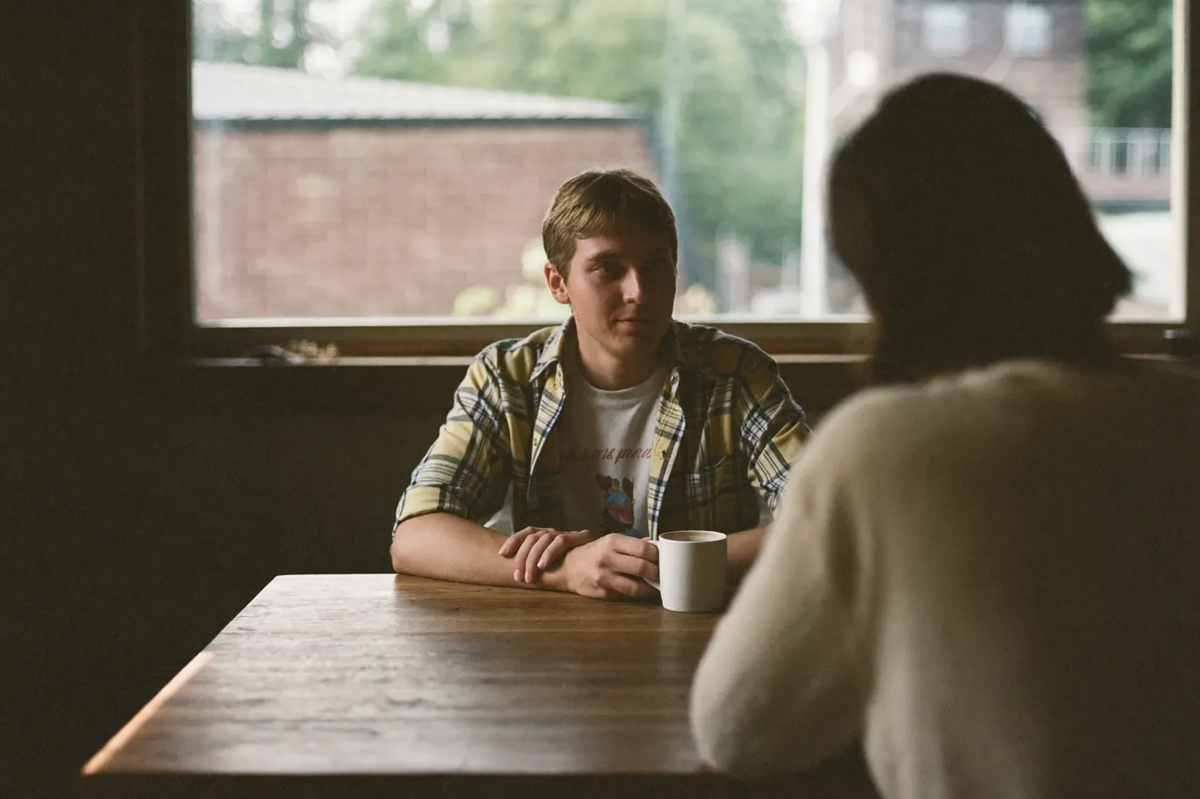 Two people in relaxed conversation over coffee at a table by a window