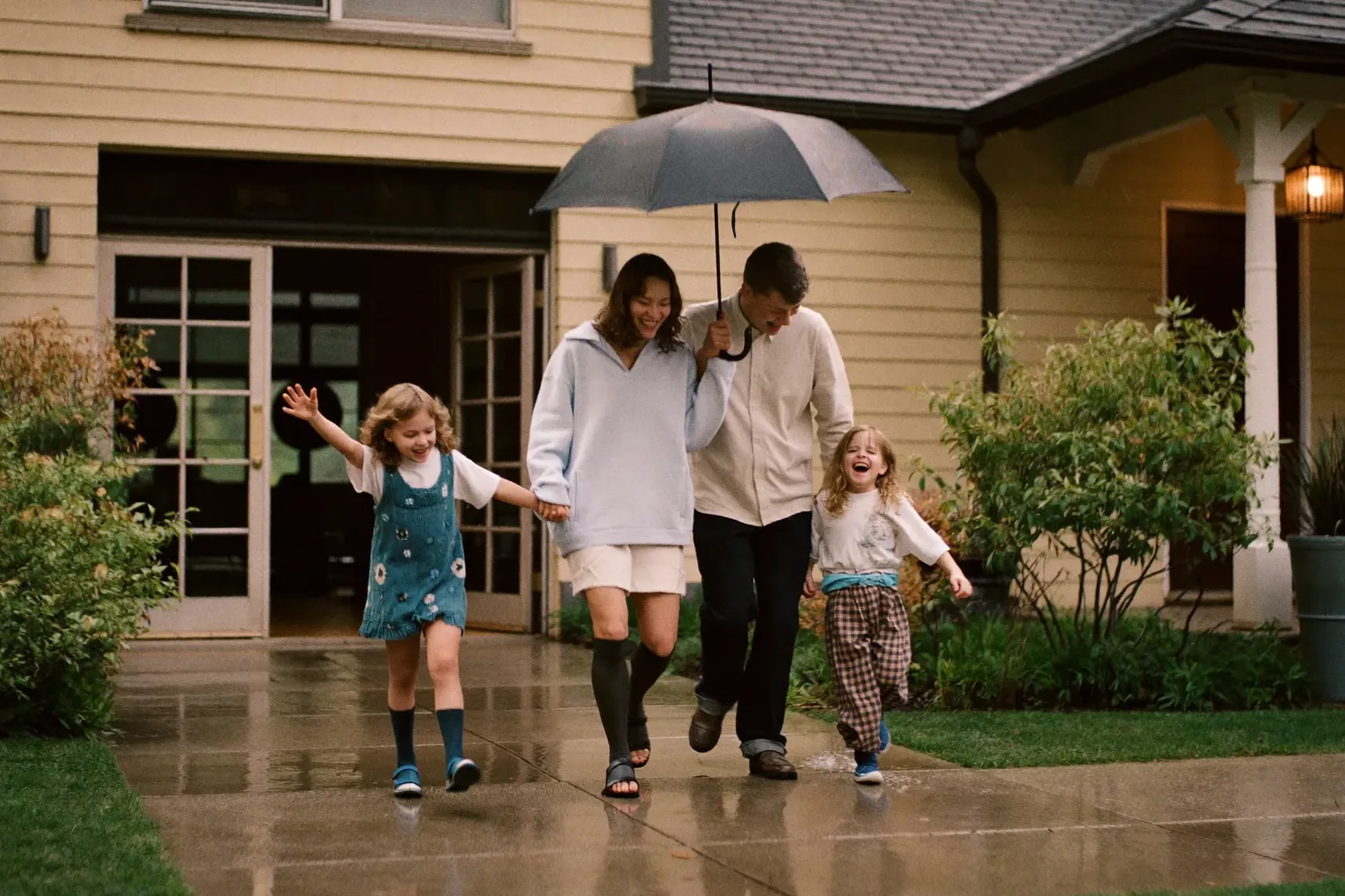 A family of four sharing one umbrella, laughing in light rain outside their home